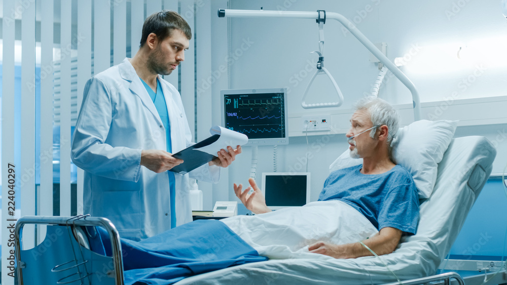 In the Hospital, Recovering Senior Patient Lying in Bed Talks with a Friendly Doctor. Modern Hospital Ward where People Get Best Health Care. In the Hospital, Recovering Senior Patient Lying in Bed Talks with a Friendly Doctor. Modern Hospital Ward where People Get Best Health Care.