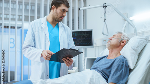 In the Hospital, Recovering Senior Patient Lying in Bed Talks with a Friendly Doctor. Professional Doctor Asks Patient Vital Questions in the Modern Geriatrics Ward.