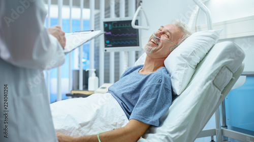 In the Hospital  Recovering Smiling Senior Patient Lying in Bed Talks with a Friendly Doctor. Professional Doctor Asks Patient Vital Questions in the Modern Geriatrics Ward.