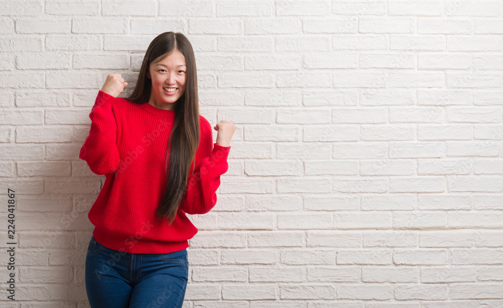 Young Chinese woman over brick wall very happy and excited doing winner gesture with arms raised, smiling and screaming for success. Celebration concept. Young Chinese woman over brick wall very happy and excited doing winner gesture with arms raised, smiling and screaming for success. Celebration concept.