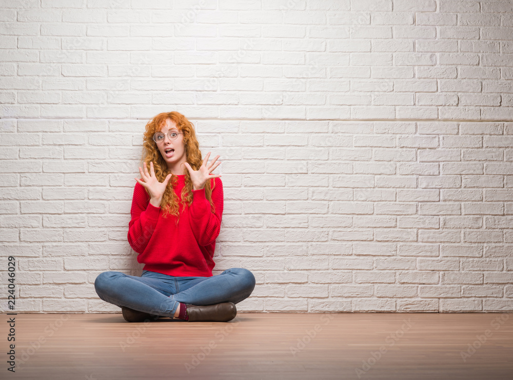Young redhead woman sitting over brick wall afraid and terrified with fear expression stop gesture with hands, shouting in shock. Panic concept. Young redhead woman sitting over brick wall afraid and terrified with fear expression stop gesture with hands, shouting in shock. Panic concept.