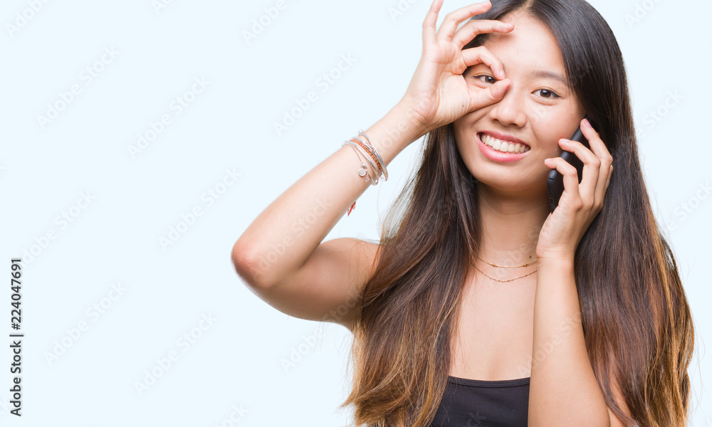 Young asian woman speaking on the phone over isolated background with happy face smiling doing ok sign with hand on eye looking through fingers Young asian woman speaking on the phone over isolated background with happy face smiling doing ok sign with hand on eye looking through fingers