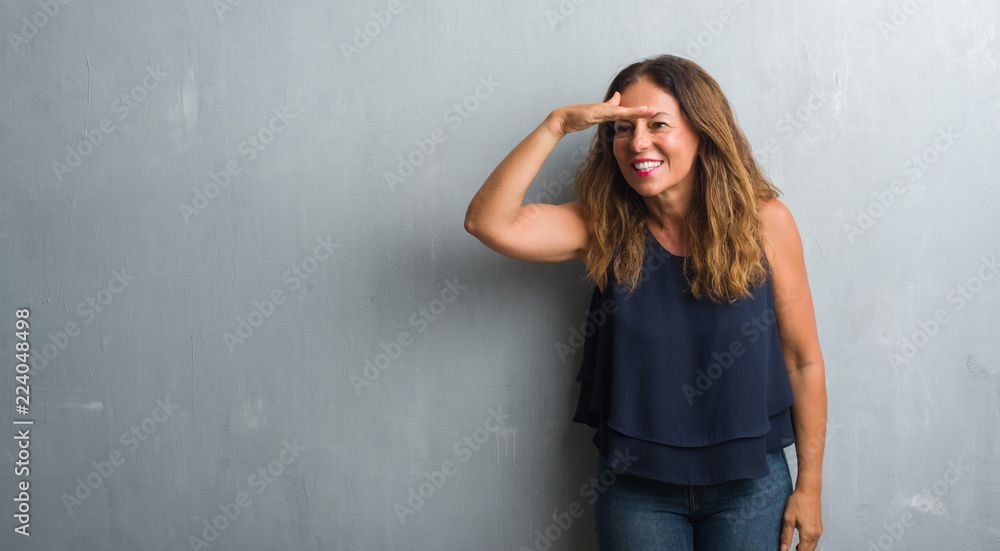 Middle age hispanic woman standing over grey grunge wall very happy and smiling looking far away with hand over head. Searching concept. Middle age hispanic woman standing over grey grunge wall very happy and smiling looking far away with hand over head. Searching concept.