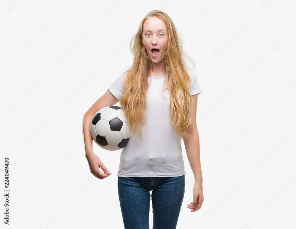 Blonde teenager woman holding soccer football ball scared in shock with a surprise face, afraid and excited with fear expression