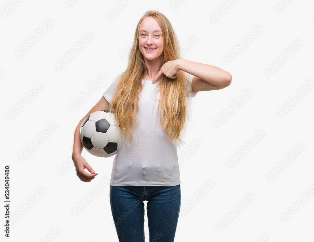 Blonde teenager woman holding soccer football ball with surprise face pointing finger to himself