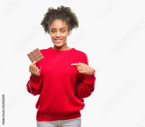 Young afro american woman eating chocolate bar over isolated background with surprise face pointing finger to himself