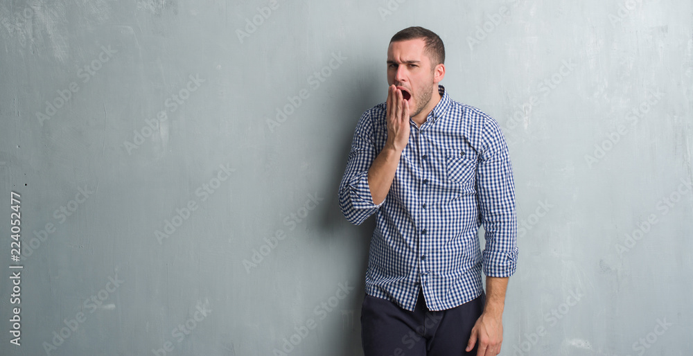Young caucasian man over grey grunge wall bored yawning tired covering mouth with hand. Restless and sleepiness.