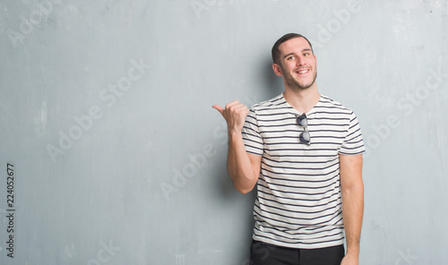 Young caucasian man over grey grunge wall smiling with happy face looking and pointing to the side with thumb up. © Krakenimages.com