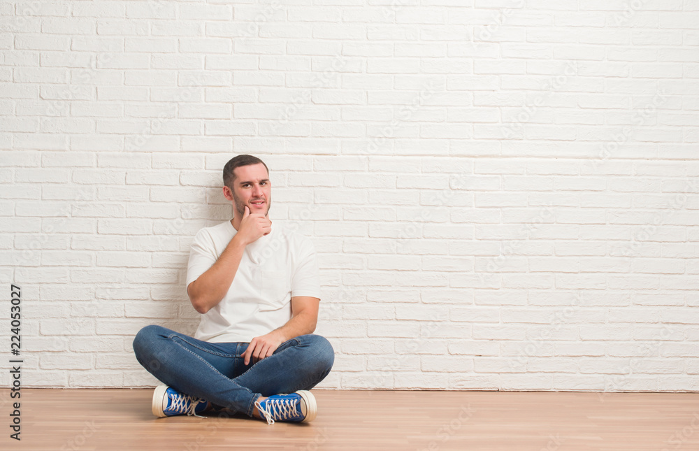 © Krakenimages.com - Young caucasian man sitting on the floor over white brick wall looking confident at the camera with smile with crossed arms and hand raised on chin. Thinking positive. © Krakenimages.com - Young caucasian man sitting on the floor over white brick wall looking confident at the camera with smile with crossed arms and hand raised on chin. Thinking positive.