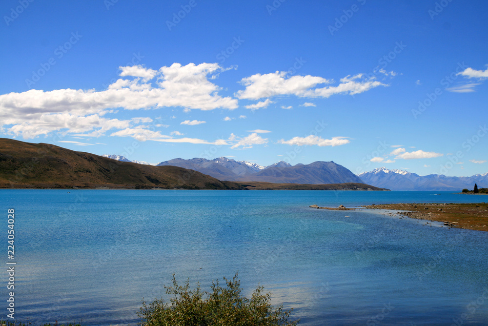 Turquoise blue lake Tekapo surrounded by hills with dry bushes and Southern Alps in back, sunny autumn day at South Island of New Zealand, Mackenzie country