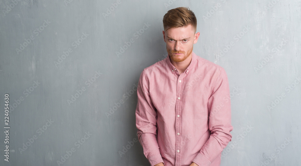 Young redhead man over grey grunge wall wearing pink shirt skeptic and nervous, frowning upset because of problem. Negative person.