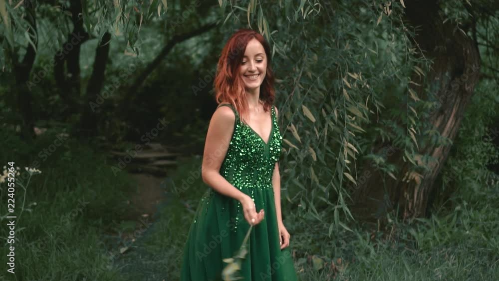 A red-haired, funny, laughing girl in a green, emerald evening dress, twists and waves a rose. Background trees, forest. Positive emotions with a smile. Creative colors A red-haired, funny, laughing girl in a green, emerald evening dress, twists and waves a rose. Background trees, forest. Positive emotions with a smile. Creative colors