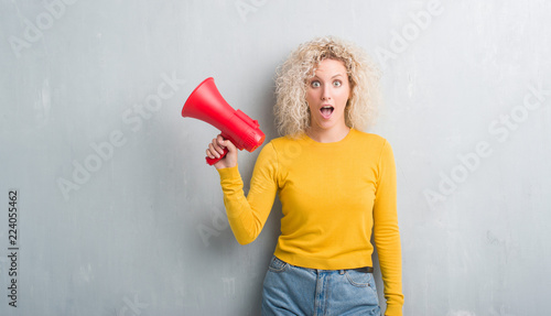Young blonde woman over grunge grey background holding megaphone scared in shock with a surprise face, afraid and excited with fear expression