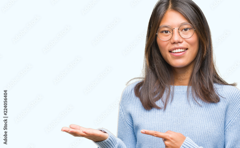 Young asian woman wearing winter sweater over isolated background amazed and smiling to the camera while presenting with hand and pointing with finger. Young asian woman wearing winter sweater over isolated background amazed and smiling to the camera while presenting with hand and pointing with finger.