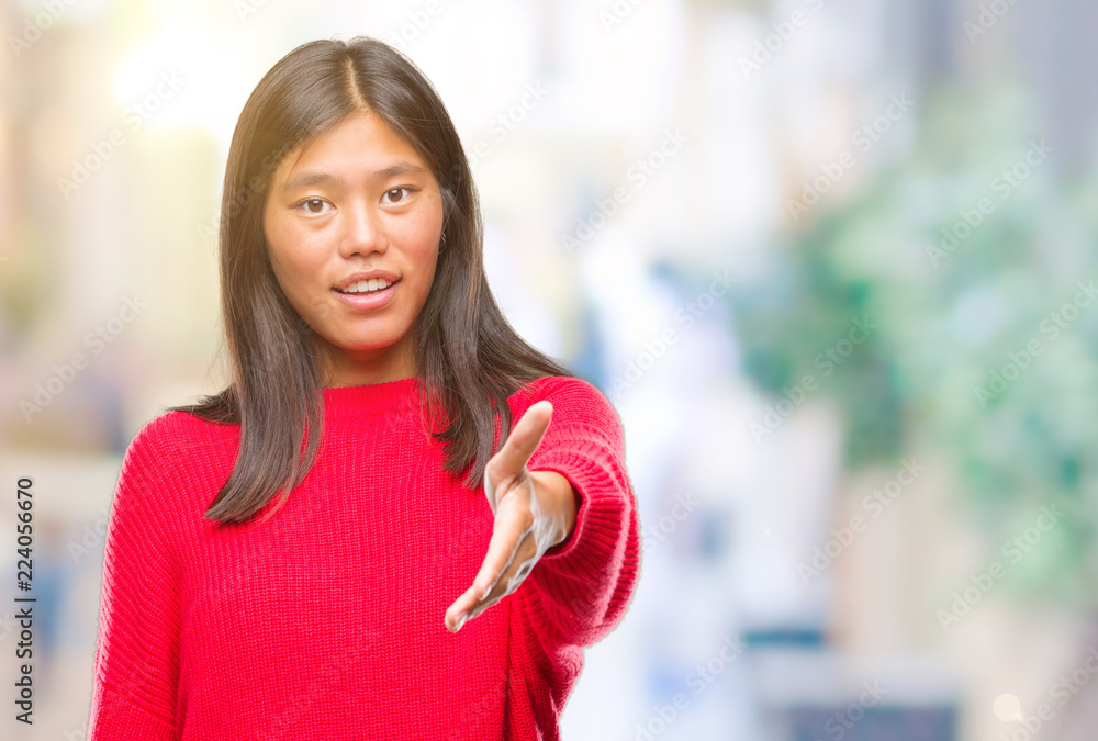 Young asian woman wearing winter sweater over isolated background smiling friendly offering handshake as greeting and welcoming. Successful business.