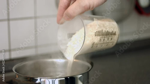 Cooking rice. Close up of a pot of boiling water, someone pouring in a beaker full of rice and stirring it with a kitchen spoon.