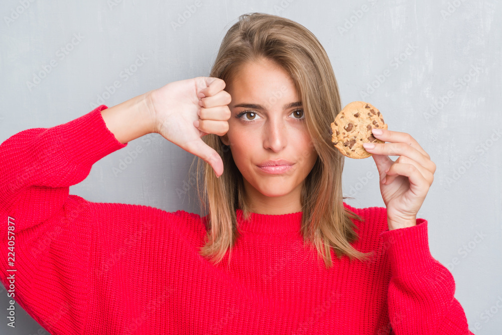 Beautiful young woman over grunge grey wall eating chocolate chip cooky with angry face, negative sign showing dislike with thumbs down, rejection concept Beautiful young woman over grunge grey wall eating chocolate chip cooky with angry face, negative sign showing dislike with thumbs down, rejection concept