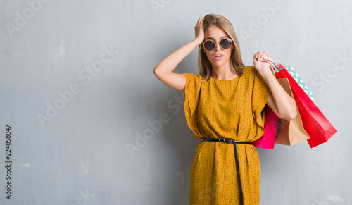Beautiful young woman over grunge grey wall holding shopping bags on sales stressed with hand on head, shocked with shame and surprise face, angry and frustrated. Fear and upset for mistake.