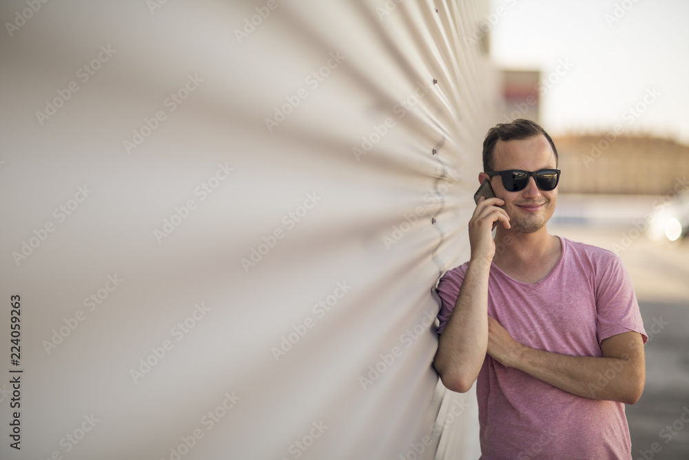 Portrait of young handsome smiling man standing next to corrugated iron wall with modern designed smartphone in hand. Nice summer sunny weather and warm colors photo. Portrait of young handsome smiling man standing next to corrugated iron wall with modern designed smartphone in hand. Nice summer sunny weather and warm colors photo.