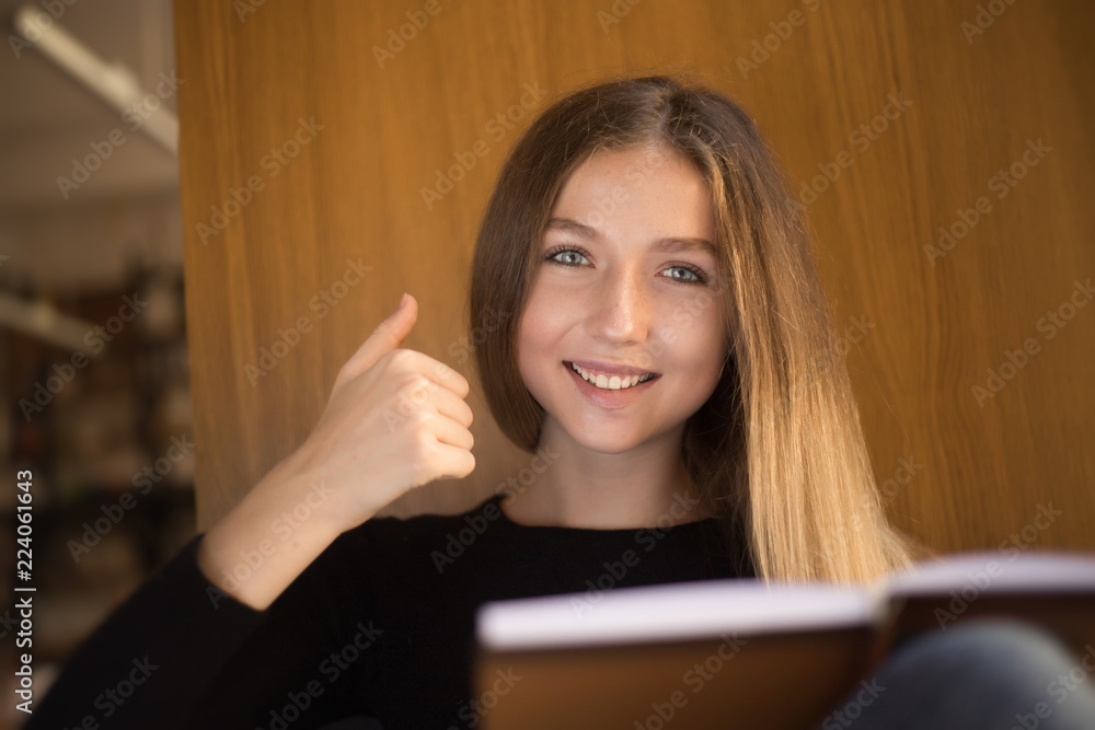 Happy young teenage girl student sitting in library reading book near window looking camera showing thumbs up gesture. Happy young teenage girl student sitting in library reading book near window looking camera showing thumbs up gesture.