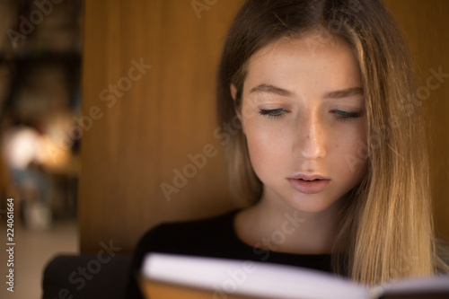 Concentrated serious young teenage girl student sitting in library reading book near window.