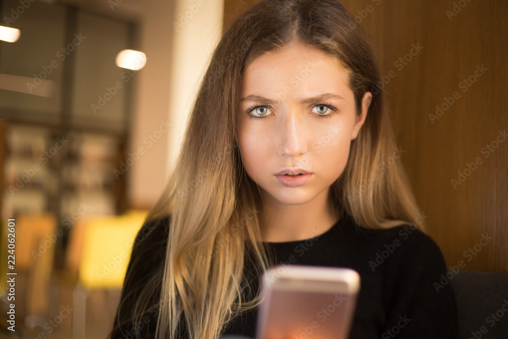 Displeased serious young teenage girl student sitting in library using mobile phone chatting near window. Looking camera. Displeased serious young teenage girl student sitting in library using mobile phone chatting near window. Looking camera.