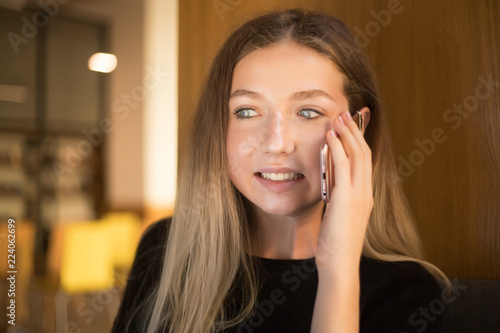 Confused funny young teenage girl student sitting in library talking by mobile phone near window.