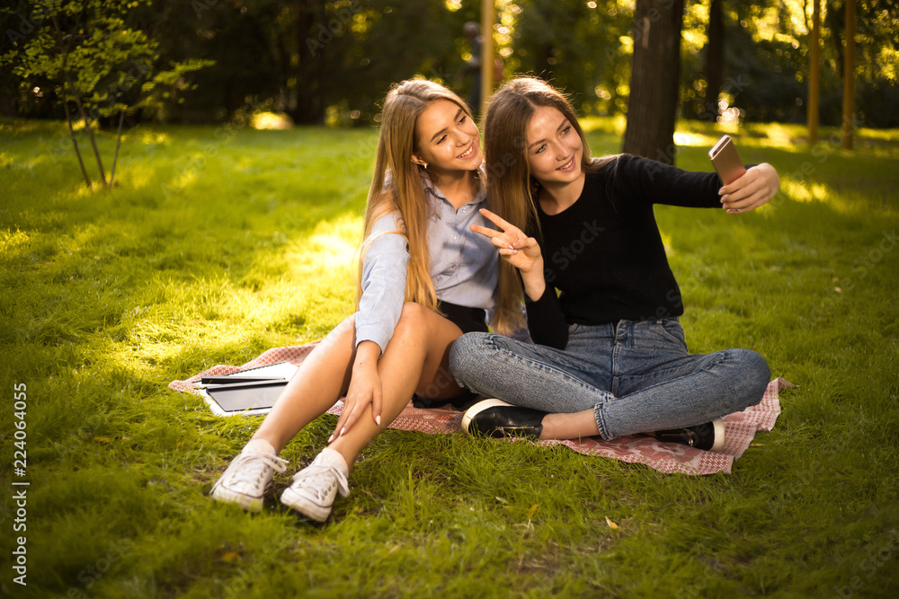 Happy girls sisters students sitting in the park outdoors on grass have a rest take a selfie by mobile phone showing peace gesture. Happy girls sisters students sitting in the park outdoors on grass have a rest take a selfie by mobile phone showing peace gesture.