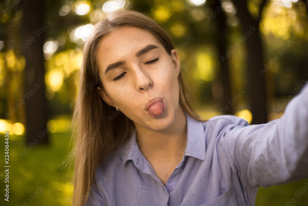 © Sunrise Team - Happy funny girl student sitting in the park outdoors on grass have a rest take a selfie by camera showing tongue.