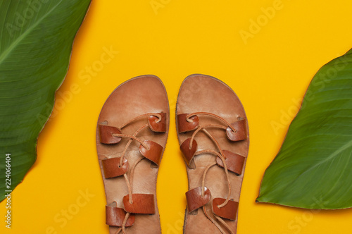 Fashionable handmade natural organic rattan bag, leather sandals, tropical leaves on bright yellow minimalistic background. Ladies bag made of natural material. Copy space, top view.  photo