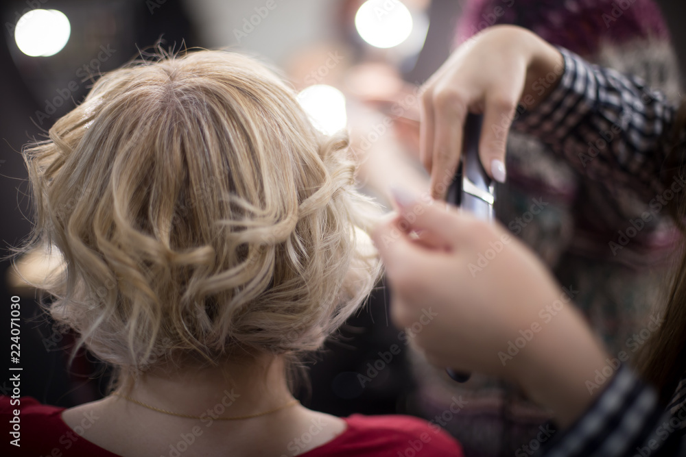 Fototapeta premium Curling tongs in the hands of a woman. Female hairdresser standing and making hairstyle to cute lovely young woman in beauty salon
