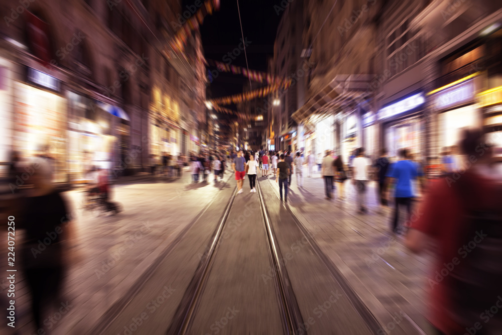 Blurry motion image of people walking in Istiklal Avenue (the city’s main pedestrian boulevard) at night in Istanbul. The street which is lined with 19th-century buildings, shopping chains and cafes.