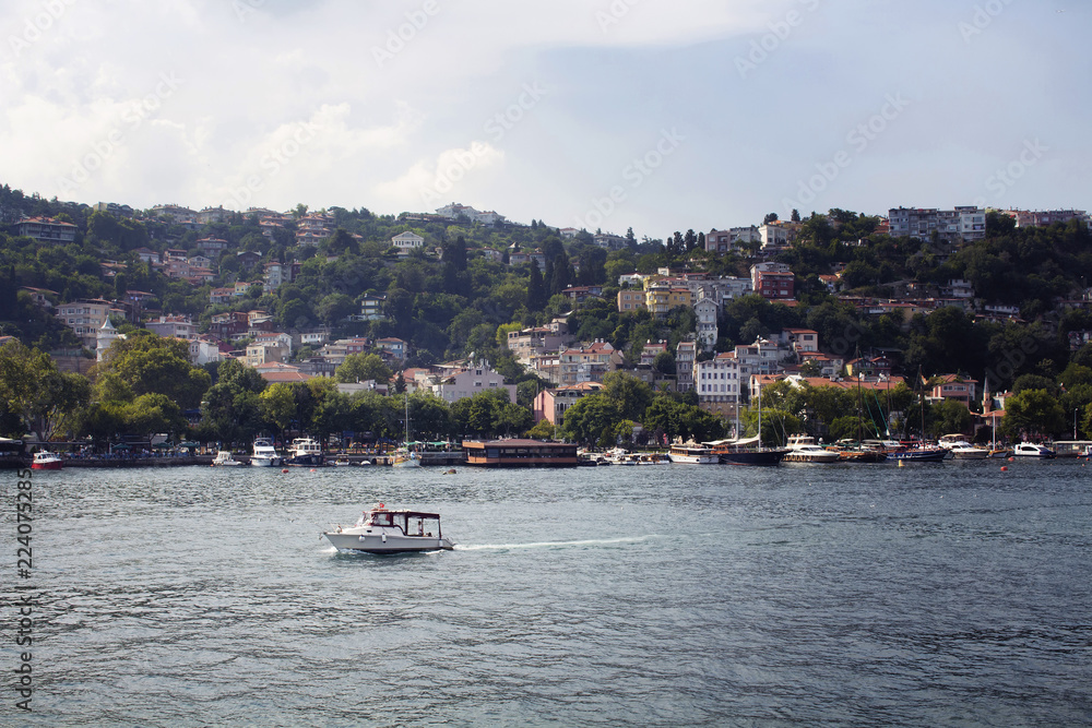 View of small fishing boat passing Bosphorus, motorboats and yachts, buildings in Kurucesme neighborhood on European side of Istanbul. It is a sunny summer day.