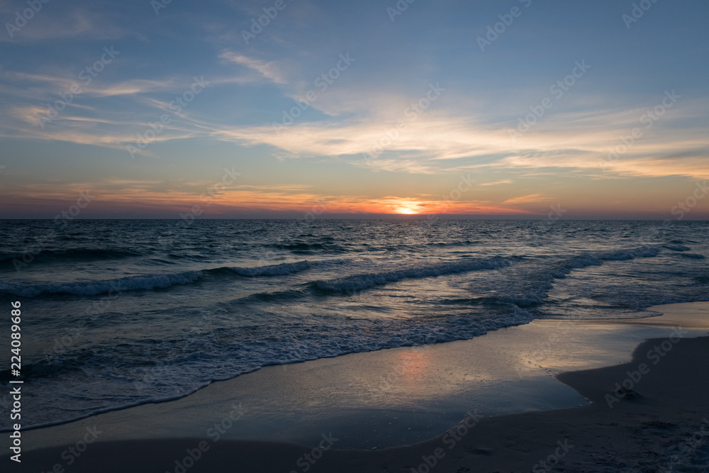 An extravagant sunset over a Florida beach in Panama City turns the sky different shades of yellow, orange, blue, green, and purple surrounded by white but soon dark clouds and blue ocean waves below.