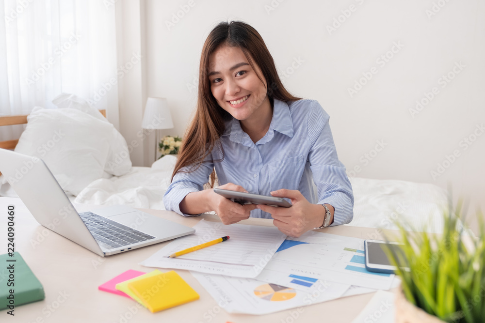 Asian freelancer looking at camera and holding tablet while working with laptop on table in bedroom at home.Work at home concept.work from home concept. Asian freelancer looking at camera and holding tablet while working with laptop on table in bedroom at home.Work at home concept.work from home concept.