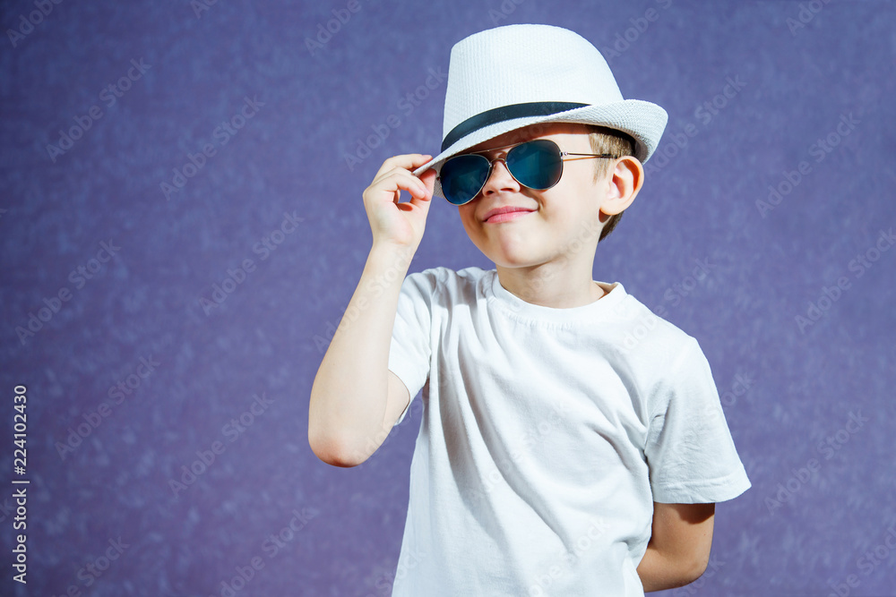 A little boy in a white hat and t-shirt in sunglasses on a purple background. The child smiles and rejoices. A little boy in a white hat and t-shirt in sunglasses on a purple background. The child smiles and rejoices.