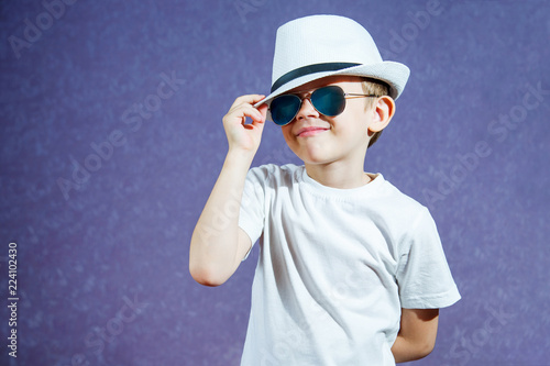 A little boy in a white hat and t-shirt in sunglasses on a purple background. The child smiles and rejoices.