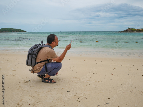 Asian fat backpacker Sitting while see his mobile phone on the idyllic beach with beautiful Sky in vacation time.Samae San Island thailand.Summer concept. © Sumeth