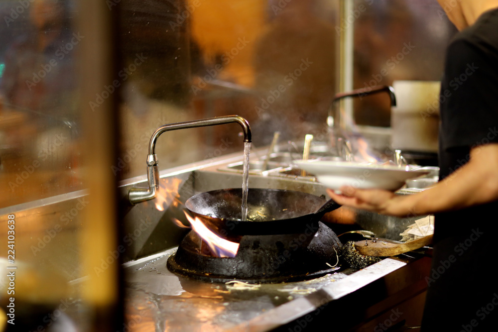 The cook in the kitchen pours a wok frying pan, standing on a special plate with an open flame The cook in the kitchen pours a wok frying pan, standing on a special plate with an open flame