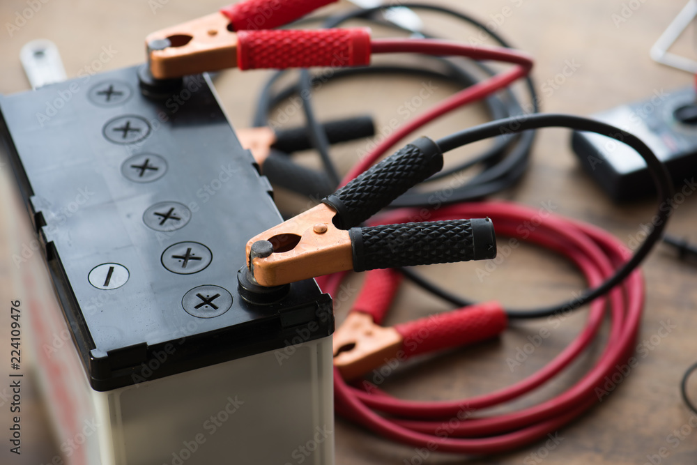 A Car battery with red and black battery Jumper Cables with copper clamps  attached to the terminals. Automotive battery on a work desk with tools in  background. Stock Photo | Adobe Stock