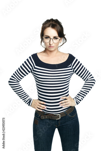 Young woman with glasses in a striped tight blouse keeping hands on waist and looks at the camera displeased.