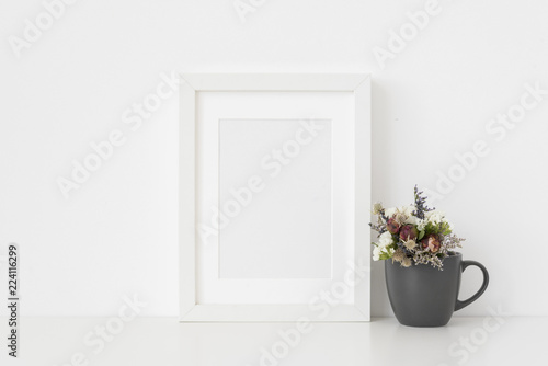 Elegant white A5 portrait frame mockup with small bouquet of dried flowers in a mug on white wall background. Empty frame  poster mock up for presentation design.