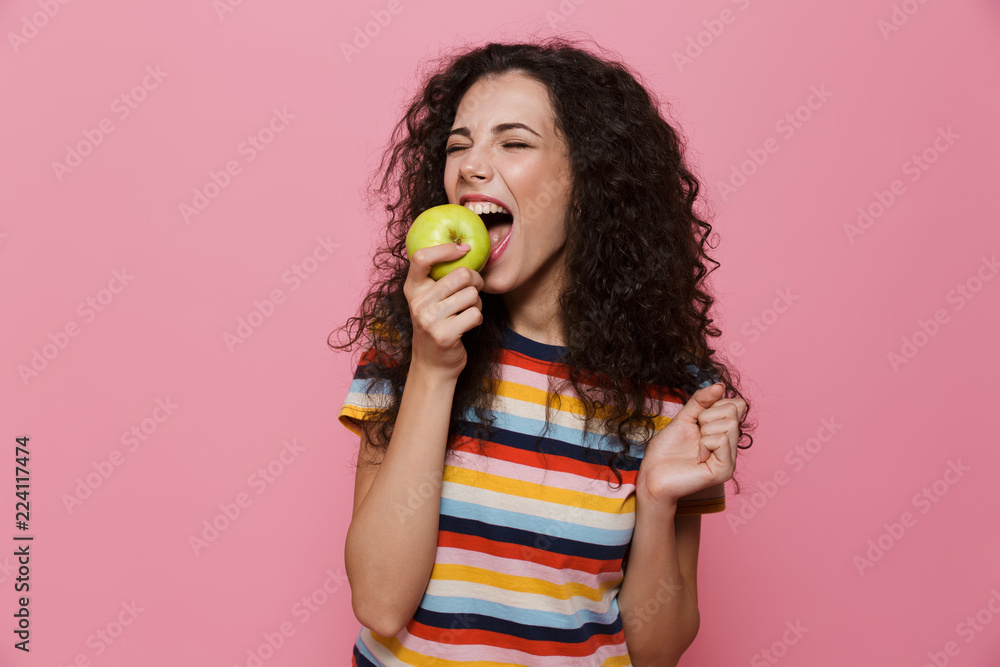 Obraz premium Photo of beautiful woman 20s with curly hair eating green apple, isolated over pink background