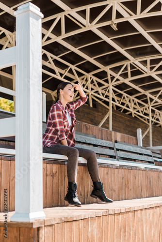 attractive cowgirl in checkered shirt sitting on bench at ranch stadium and looking at something