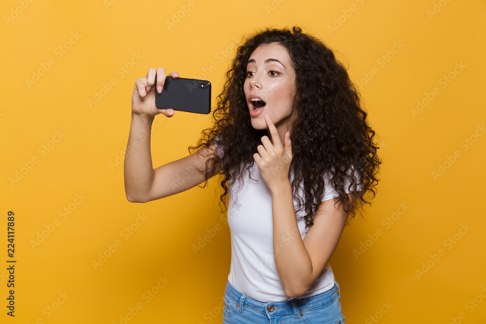 Image of attractive woman 20s with curly hair laughing and taking selfie photo on mobile phone, isolated over yellow background Image of attractive woman 20s with curly hair laughing and taking selfie photo on mobile phone, isolated over yellow background
