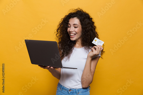 Photo of content woman 20s wearing casual clothes holding black laptop and credit card, isolated over yellow background