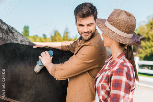 Fototapeta handsome male equestrian cleaning black horse with brush at ranch and looking at