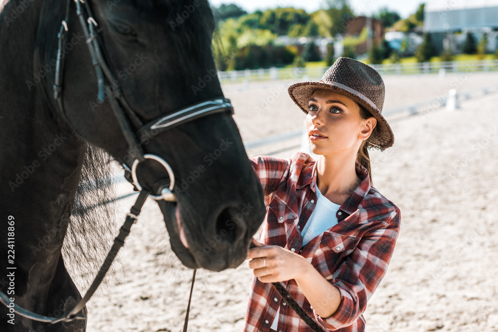 Obraz premium attractive jockey in checkered shirt and hat fixing horse halter at ranch