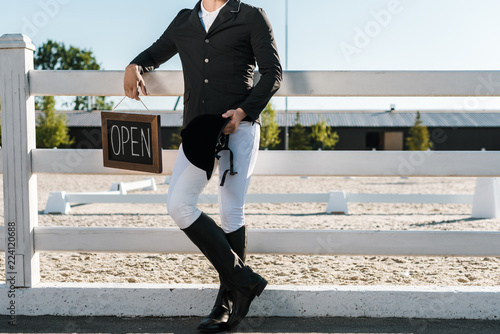 cropped image of male equestrian leaning on fence and holding open sign at horse club