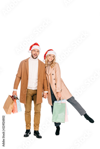 beautiful happy young couple in santa hats holding shopping bags and smiling at camera isolated on white